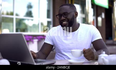 Smiling freelancer working on laptop drinking coffee and looking at camera Stock Photo
