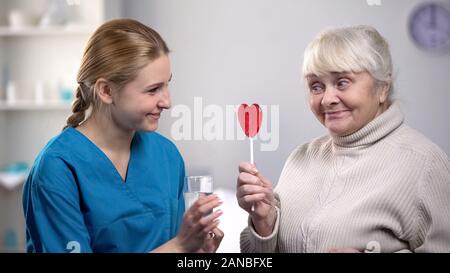 Nice volunteer giving glass of water to funny elderly woman holding lollipop Stock Photo
