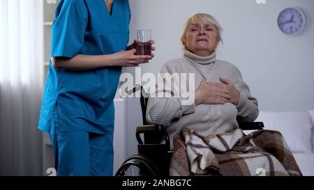 Female nurse giving pills and water glass old lay suffering chest pain, medicine Stock Photo