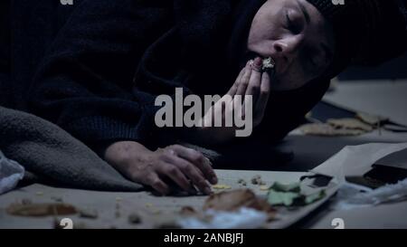 Hungry homeless person lying on ground with garbage and greedily eating orts Stock Photo