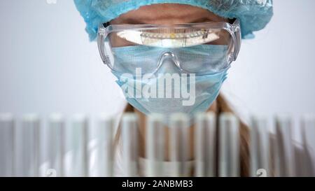 Female chemist in protective mask looking at test tubes, biological research Stock Photo