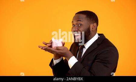 Cheerful afro-american man in suit smiling and holding piggybank, trustful bank Stock Photo