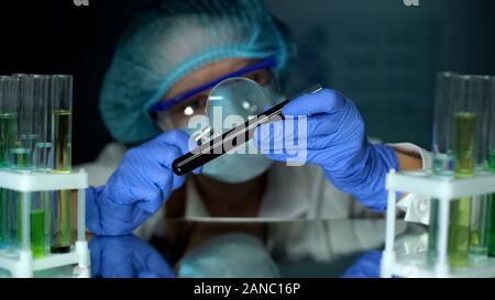 Scientist looking at black substance in tube with magnifying glass, examination Stock Photo