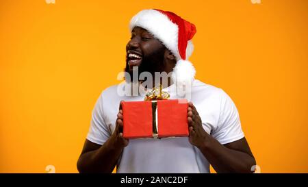 African-American man in Santa hat presenting Christmas gift, yellow background Stock Photo
