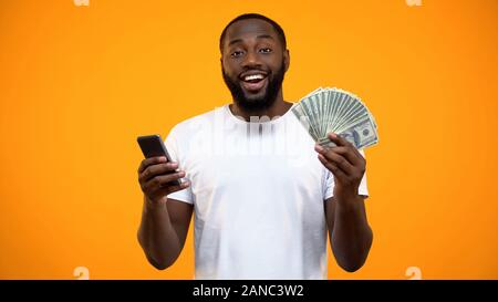 Excited Afro-American man holding smartphone and dollars, online money transfer Stock Photo
