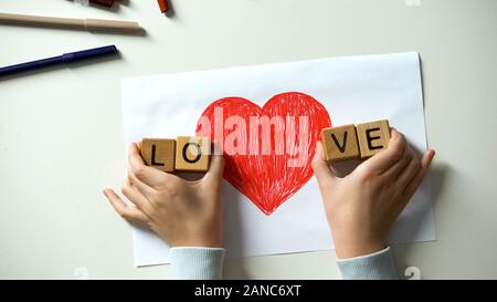 Kid putting love word made of wooden cubes on sides of red heart drawing Stock Photo