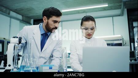 Pretty young Caucasian woman in glasses working in the laboratory and doing some test in the tube with liquid, then tapping on the laptop computer. Stock Photo