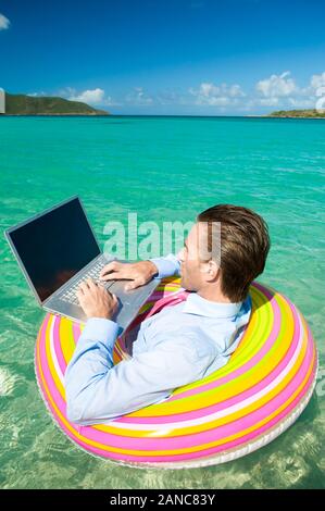 Relaxed businessman floating on a rubber ring while typing on his laptop Stock Photo