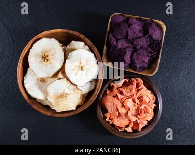 Dried carrot, beetroot and apple chips in a wooden bowl on a black stone background. Organic natural food. Top view, flat lay. Stock Photo