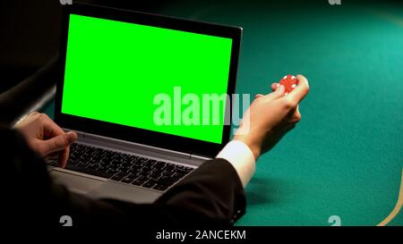 Man using online betting services on laptop, holding lucky chip, green screen Stock Photo