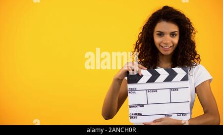 Smiling young female holding clapper board on orange background, cinematography Stock Photo