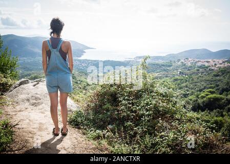 Young woman admiring panorama from hill overseeing bay in Tuscany wearing denim dungaree Stock Photo