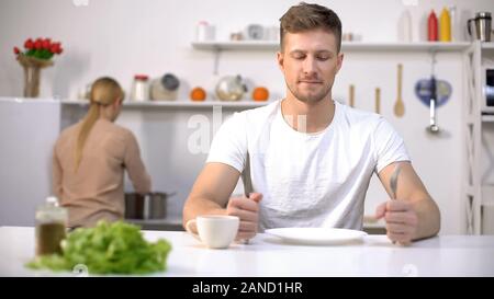 Positive man waiting for dinner, young wife cooking soup on background, hunger Stock Photo