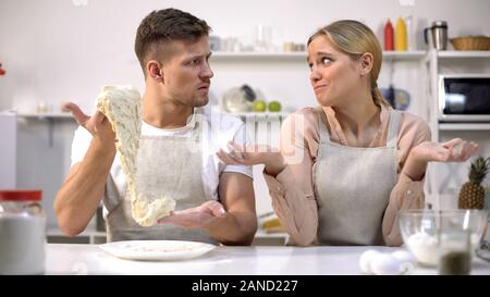 Clumsy man stretching dough, looking at wife, difficulties during cooking Stock Photo