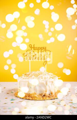 Birthday cake with golden topper. Birthday party celebration concept. Vertical, bold yellow background, bright lights bokeh background Stock Photo