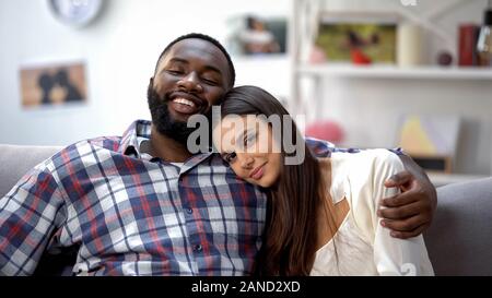 Smiling multiethnic couple hugging on sofa, relaxing together, tender feelings Stock Photo
