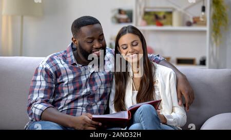 Cheerful mixed-race couple viewing family photo album at home, pleasure memories Stock Photo