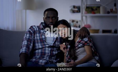 Emotional mixed-race couple with pop corn watching horror movie, evening at home Stock Photo