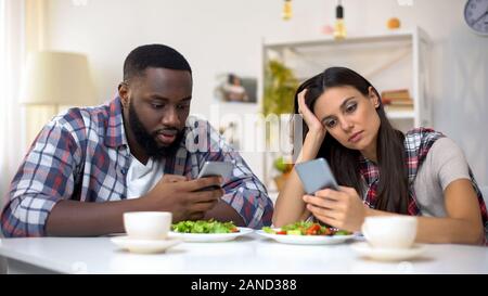 Offended mixed-race couple using smartphones during lunch, misunderstanding Stock Photo