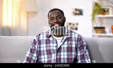 Cheerful African-American man sitting on sofa and looking at camera, relaxing Stock Photo