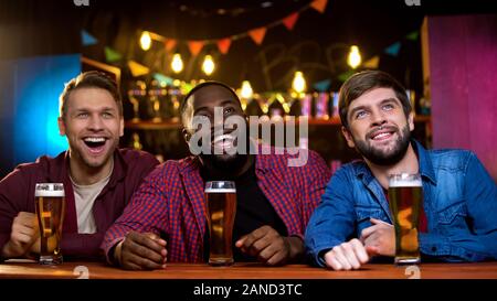 Multi-ethnic friends smiling watching comedy show on big screen in beer pub, fun Stock Photo
