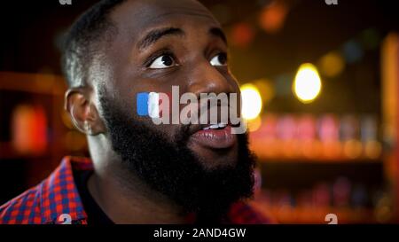 Emotional black man with hope in eyes and french flag on cheek watching contest Stock Photo