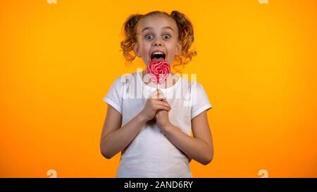 Pretty girl licking delicious heart-shaped lollipop, childhood, confectionery