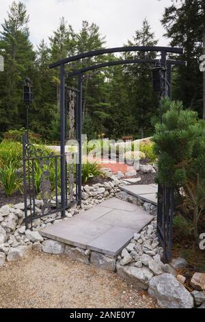 Grey paving tile path through black wrought iron metal arbour leading to borders with mixed plantings in front yard country garden in summer. Stock Photo