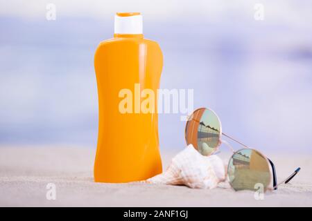 Close-up of an orange plastic bottle next to a pair of sunglasses resting on a seashell on the sand on an out of focus background. Summer vacation con Stock Photo