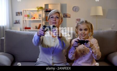 Cute girl and modern granny playing video game at home with joysticks, leisure Stock Photo