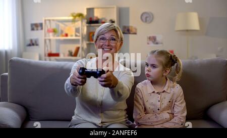 Excited granny playing video game ignoring sad grandchild sitting sofa addiction Stock Photo