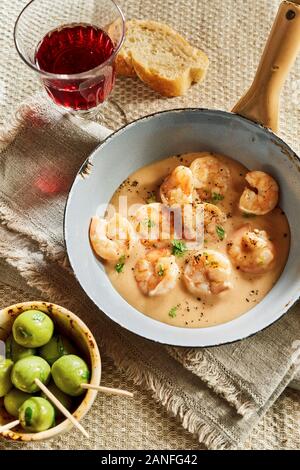 Top view of skampi lobsters in cheese cream sauce, served on rustic table in small pan with wooden handle, bowl of green olives, white bread and a gla Stock Photo