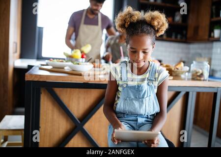 Cute african american girl using a tablet while her parents preparing food in kitchen