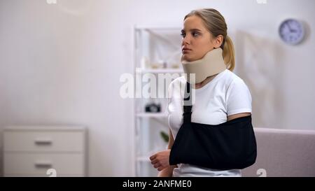 Unhappy woman in foam cervical collar and arm sling sitting on sofa, rehab Stock Photo