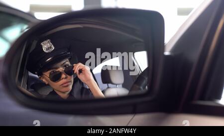 Confident policewoman putting on sunglasses looking into rearview mirror of car Stock Photo
