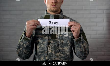 Pray word written on sign in hands of male soldier, serviceman asking for peace Stock Photo