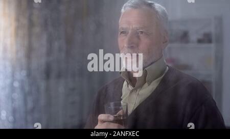 Retired male enjoying taste of whisky home, looking outside through rainy window Stock Photo