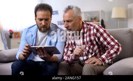 Adult son and senior father watching tutorial videos on tablet, information Stock Photo
