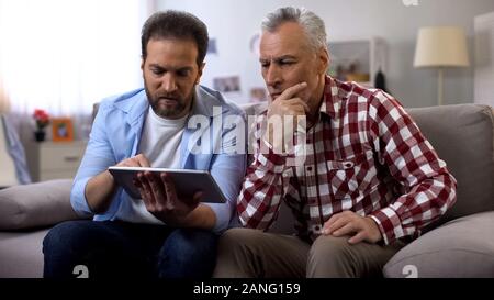 Middle-aged son teaching retiree dad to use banking app on tablet, technologies Stock Photo