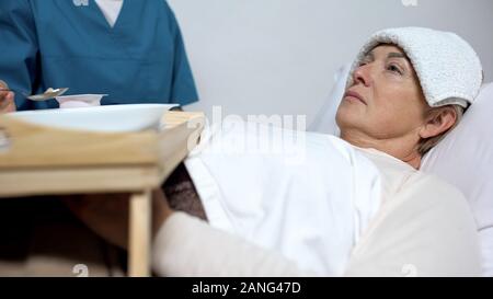 Nurse feeding sick indifferent woman with compress on head, lying in hospice bed Stock Photo