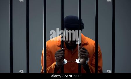 Upset afro-american criminal in handcuffs holding prison bars, regretting past Stock Photo