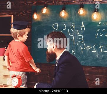 Teacher and child talking in the classroom. Little schoolboy in Oxford cap answering the question in front of the green board. Male adult sitting at t Stock Photo