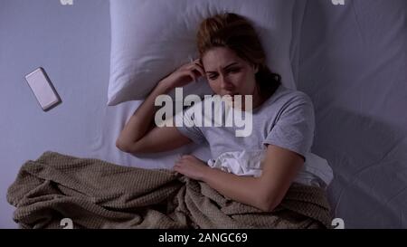 Depressed woman lying in bed covered in blanket with smartphone beside, solitude Stock Photo