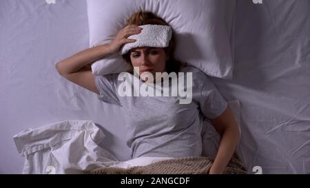 Upset woman with compress lying in bed, suffering from migraine and nausea Stock Photo