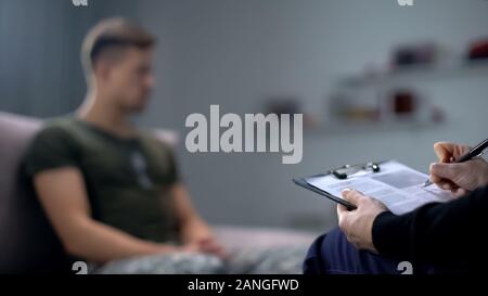 Psychologist making notes during therapy session with sad male soldier, PTSD Stock Photo
