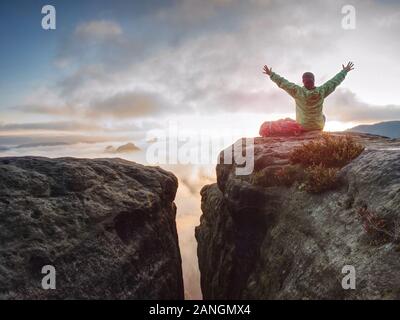 Young woman sit down on top of a mountain celebrate the day. Young lady hiker sitting in blue green windbreaker with red rucksack on sharp cliff Stock Photo
