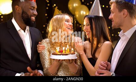 Cheerful female holding birthday cake preparing to blow candles, friends around Stock Photo