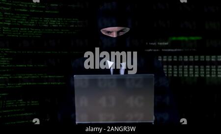 Hacker in balaclava working on laptop, illegal privacy attack, data leakage Stock Photo