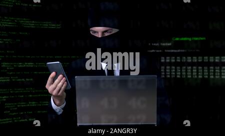 Criminal in suit and balaclava breaking security code using laptop and phone Stock Photo