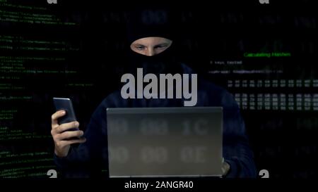 Criminal in mask checking surveillance camera on laptop and phone, database Stock Photo